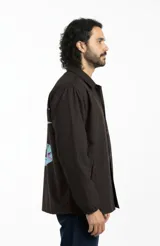 Campera impermeable marrón con estampa en la espalda de un ser orbitando un anillo con tipografía glitch. Corte relajado estilo coach, cuello camisero, broches a presión al frente, bolsillos laterales y puños elastizados.