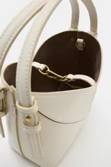 Bolso tipo mini shopper de diseño estructurado en color beige, con forma de V en la parte superior y asa de mano fija. Presenta costuras visibles y detalles metálicos en los laterales.