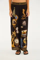 Pantalón largo de silueta amplia, color negro, con diseño estampado de diversos objetos como relojes, palmeras, animales y marcos estilo barroco. Cuenta con cintura elastizada y detalles decorativos en los bordes de la cintura y los tobillos.