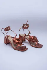 Sandalias de cuero color cobre con taco cuadrado y tira fina sobre los dedos. Presentan una flor grande en el empeine y tiras finas para atar al tobillo.