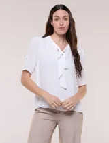Blusa blanca de corte recto y tejido liviano de gasa con forro interno delantero. Presenta escote en V con cuello redondo y lazo central para anudar, mangas cortas con hilera de botones forrados en los laterales y ruedo ligeramente redondeado.