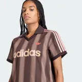 Camiseta de fútbol Adidas de manga corta, color marrón con diseño de rayas verticales oscuras. Presenta cuello polo con escote en V, el logo clásico de la marca bordado en el pecho junto con la inscripción "adidas" en gran tamaño al frente, y las franjas características de la marca en los hombros.