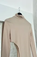 Top de lycra color beige, con cuello alto y mangas largas. Presenta un corte asimétrico en la parte inferior.