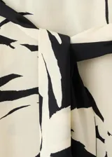 Mono largo de viscosa con diseño bicolor en negro y beige con estampado de hojas tropicales. Presenta un corte asimétrico de un solo hombro con manga larga holgada, pantalón de silueta recta y lazo ajustable en la cintura.