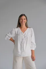 Blusa blanca de corte holgado con cuello en V, cierre frontal con botones y detalles de alforzas y encaje en el pecho. Presenta mangas largas con puños rectos.