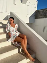 Vestido corto blanco con estampado floral en tonos naranja y verde, escote en V con detalle de nudo, mangas largas acampanadas y falda con volantes y abertura lateral.