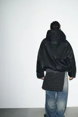 Chaqueta acolchada de color negro con acabado repelente al agua y resistente al viento. Presenta cuello subido con capucha ajustable, cierre frontal con cremallera termosellada, bolsillos delanteros con cremallera y bajo con ajuste elástico. Las mangas largas cuentan con tiras adhesivas ajustables en los puños.