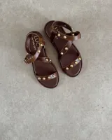 Sandalias planas de cuero negro con tiras decoradas con tachas metálicas doradas. Presentan una tira frontal sobre los dedos y una tira ajustable con hebilla alrededor del tobillo.