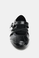 Zapato tipo bailarina de acabado acharolado en color negro, con punta redonda y suela plana. Presenta una tira ajustable con hebilla metálica y bandas elásticas cruzadas en el empeine.