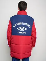 Parka larga de abrigo Umbro del Club Nacional de Football, con diseño tricolor en rojo, azul y blanco. Presenta cuello alto, cierre frontal con solapa, bolsillos con cierre en el pecho y logo de la marca junto al escudo del club.