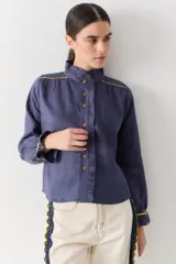 Camisa de lino color azul, con cuello alto tipo mao, cierre frontal con botones y detalle de volados en la cartera. Presenta mangas largas con puños y vivos en color amarillo a contratono en hombros y puños.