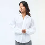 Camisa de popelina blanca con cuello clásico, cierre frontal de botones y mangas largas abullonadas con puños ajustados.