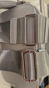 Cartera tipo bandolera de diseño rectangular y estructura rígida, color gris. Cuenta con doble compartimento superior con cierre metálico, herrajes plateados y correa de hombro ancha, ajustable y extraíble.