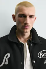 Cazadora estilo varsity con cuerpo de tejido con lana y mangas de efecto piel. Presenta cuello solapa, cierre frontal con botones a presión, bolsillos laterales y puños con elástico a rayas. Incluye parches y bordados decorativos en el frente.