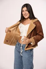 Campera tipo gamulán de gamuza color tostado con interior y solapas de corderito sintético color crema. Presenta cierre frontal con botones, bolsillos cuadrados delanteros y detalles de costuras visibles.