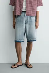 Bermuda de denim de algodón con efecto lavado desgastado en tonos marrones y azules. Tiene tiro medio, diseño de cinco bolsillos, pierna recta de corte holgado y bajo con acabado irregular. Cuenta con cierre frontal de cremallera y botón metálico.