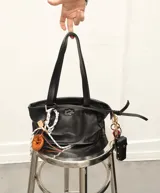 Charm para bolso con forma de pretzel trenzado en cuero color caramelo, sujeto con cordón blanco y negro trenzado. Incluye un pequeño dije rectangular de madera con grabado.