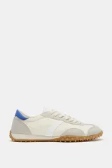 Zapatilla deportiva estilo blucher de color blanco, con detalles en gris claro, una franja lateral blanca y una talonera azul. Cuenta con cordones, punta redonda y suela de goma con tachuelas.