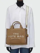 Bolso tipo tote bag de Marc Jacobs en color beige, confeccionado en lona resistente. Presenta el texto "THE TOTE BAG" y "MARC JACOBS" impreso en la parte frontal en color negro. Cuenta con asas superiores y una correa de hombro ajustable y extraíble, además de ribetes laterales negros con inscripciones.