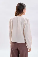 Blusa casual de color crudo con mangas largas abullonadas, cuello redondo y cierre frontal con botones. Presenta delicados detalles de encaje y pliegues en la parte frontal.