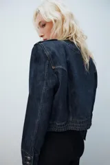 Chaqueta de denim de corte corto, color azul oscuro, con cuello redondo, hombreras y cierre frontal con cremallera. Presenta diseño de inspiración militar con doble botonadura metálica decorativa en el frente, trabillas con botón en los hombros y bajo elástico.