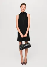 Vestido corto negro sin mangas, con diseño recto y cuello alto tipo halter con detalle de lazo. Presenta un panel frontal semitransparente en la parte superior.