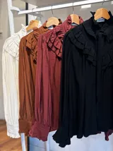 Blusa blanca de estilo romántico con volados tipo shabot en el frente, cuello alto con lazo ajustable y mangas largas abullonadas con puños ajustados.