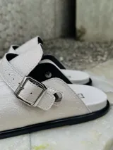 Zuecos blancos de cuero sintético con hebilla metálica ajustable.
