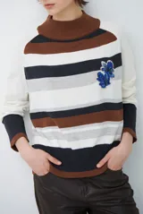 Polera de tejido de punto con diseño de rayas horizontales en tonos blanco, marrón y gris. Presenta cuello alto, mangas largas con puños acanalados en contraste y un aplique bordado de flores azules en el pecho.