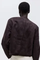 Chaqueta corta de gamuza sintética en color marrón oscuro, con forro interior de peluche. Presenta cuello redondo, cierre frontal con botones metálicos dorados y dos bolsillos delanteros con solapa y botón.