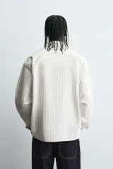 Camisa de corte relajado confeccionada en tejido de algodón, con diseño de rayas verticales. Presenta cuello solapa, cierre frontal de botones, manga larga con puños abotonados y un bolsillo plastrón en el pecho con un pequeño bordado decorativo.