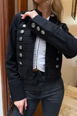Campera estilo militar de color negro, con cuello mao y cierre frontal. Presenta múltiples botones metálicos plateados dispuestos en filas horizontales y detalles de charreteras en los hombros. Las mangas cuentan con puños decorados con botones a juego.