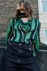 Camisa de manga larga con escote fluido tipo pañuelo, hombreras y puños abotonados. Presenta un diseño de rayas verticales en tonos verdes y amarillos.