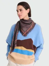 Pañuelo cuadrado tipo chal, color marrón con diseño jacquard del logo de la marca y bordes estampados en tonos naranjas y marrones, con terminación en flecos sutiles.
