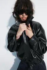Chaqueta de cuero sintético negro con cuello alto, cierre frontal de cremallera oculto por solapa con botones a presión, mangas largas y bolsillos laterales. Diseño con volumen y bajo elástico.