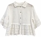 Camisa de lino blanca de corte corto y holgado, con cuello camisero y cierre frontal de botones. Presenta mangas acampanadas tres cuartos y detalles de bordado inglés en los puños y en el volado del ruedo.