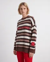 Sweater de punto oversized con diseño de rayas horizontales en tonos marrón, blanco y negro, cuello alto y mangas largas con hombros caídos. Cuenta con aberturas laterales en el dobladillo y una etiqueta roja decorativa en la manga.