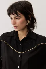 Camisa de manga larga color negro, con cuello camisero, cierre frontal con botones y detalles de ribetes contrastantes en color dorado. Presenta paneles superiores con textura y mangas abullonadas.