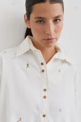 Camisa de corte holgado color blanco, con cuello clásico, cierre frontal de botones y bolsillo delantero. Presenta un delicado bordado de figuras deportivas en color marrón.