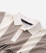 Remera tipo polo de corte oversized, confeccionada en tejido de punto con diseño de rayas verticales en tonos beige y blanco. Presenta cuello clásico, abertura frontal con botones y bordado con la inscripción 'The Hamptons New York' en el pecho.