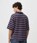 Remera polo de algodón con diseño a rayas horizontales en azul marino, rojo y blanco, corte boxy amplio, mangas cortas y cuello polo con botones. Presenta un pequeño bordado en el pecho.