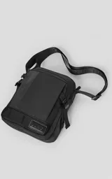 Bolso cruzado (bandolera) negro de tela sintética, con correa ajustable estampada con la palabra "LUXO". Presenta un diseño estructurado con bolsillo frontal y parche con logo en la base.