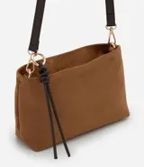 Bolso bandolera de tamaño mediano confeccionado en gamuza sintética color marrón tostado. Presenta un formato rectangular estructurado, cierre superior con cremallera y correa larga ajustable en color marrón oscuro. Incluye un detalle decorativo de tiras anudadas en uno de los laterales.