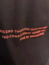 Remera negra de corte oversize con estampa en la espalda en color rojo que dice: "ASCEND THROUGH DISCIPLINE AND CONSISTENCY WHEN ONE IS WATCH".