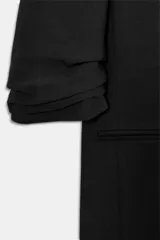 Blazer negro con cuello solapa con muescas, hombreras y forro interior a tono. Presenta manga por debajo del codo con detalle de drapeado y falsos bolsillos de vivo en el delantero, con cierre frontal de un solo botón dorado.