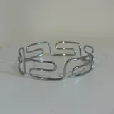 Brazalete rígido de metal plateado con diseño geométrico abstracto.
