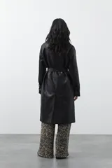 Trench coat de símil cuero color marrón oscuro, con solapas amplias, cierre frontal con botones, cinturón ajustable en la cintura y mangas con tiras ajustables. Presenta un largo por debajo de la rodilla y bolsillos laterales.