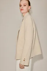 Blazer de corte estructurado en color beige, con cuello mao y cierre frontal cruzado mediante botones. Presenta mangas largas con detalle de botones en los puños y bolsillos de vivo en el frente.