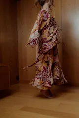 Maxi vestido estilo boho con estampado floral en tonos rosa, naranja y borgoña. Tiene mangas largas sueltas, escote redondo con volados y falda larga con volados.