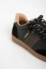 Zapatilla urbana de caña baja, con capellada combinada en tonos verde oscuro, marrón y negro. Presenta puntera y talón en gamuza negra, detalles en cuero marrón en el lateral y forro interior color beige. La suela es de goma color caramelo.